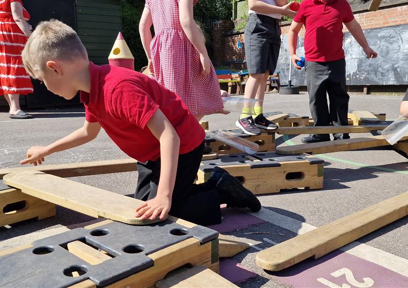 A child is placing a wooden plank onto a wooden block from the Play Builder Engineer Set. As the child is building an obstacle course, a group of children are trying to complete the course.